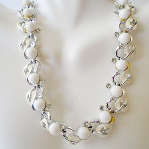 Op de afbeelding: Een witte en zilveren ketting met een kettingschakelontwerp en witte ronde kralen. De ketting is op een wit mannequin.