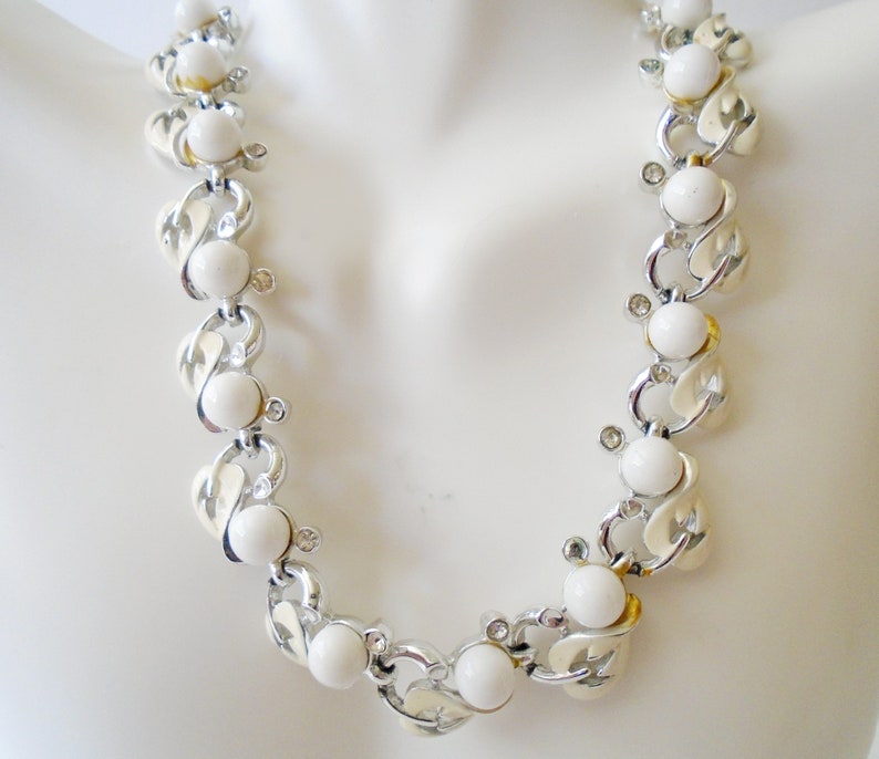 Op de afbeelding: Een witte en zilveren ketting met een kettingschakelontwerp en witte ronde kralen. De ketting is op een wit mannequin.