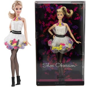May include: Barbie doll wearing a white dress with a black belt and colorful shoe-shaped embellishments. She is wearing black fishnet stockings and black boots. The doll is packaged in a box with a black background and the text "Shoe Obsession" in white. The box also features a pink label with the text "Barbie Collection Pink Label".