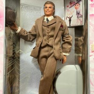 May include: A Ken doll in a brown tweed suit, holding a matching hat. The doll is in a display box with a "My Fair Lady" poster. The suit includes a jacket, waistcoat, and trousers, with a white shirt and patterned tie.