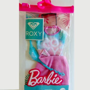 May include: A Barbie clothing set in a clear plastic package. The set includes a tie-dye top, pink skirt, blue purse, and sunglasses. The Roxy logo is visible on a teal tag. The Barbie logo is in pink and white.