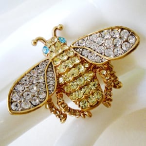 May include: A gold-toned bee brooch with clear and yellow rhinestones. The bee has blue eyes and its wings are made of clear rhinestones.