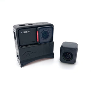 May include: A black action camera with a lens cover and the text "ONE RS" on the front. The camera is attached to a black accessory with the text "RS" on the side. A separate black lens attachment is also shown.