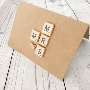 May include: A brown card with the word "MRS" spelled out using wooden letter tiles. A small wooden heart with the word "Love" is placed below the tiles.