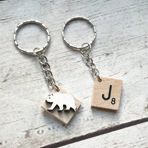 May include: Two silver-toned metal keychains with chain links. One keychain features a square light-colored wooden tile with a white bear silhouette. The other keychain features a square light-colored wooden tile with the black letter J and number 8.