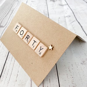 May include: A brown birthday card with the word "FORTY" spelled out in wooden Scrabble tiles. A small gold star is placed next to the word. The card is angled, revealing its folded design.