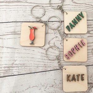 May include: Four wooden keychains with different designs. One keychain has a cocktail drink with a red umbrella, another has the name "DANNY" in green letters, another has the name "SOPHIE" in pink letters, and the last one has the name "KATE" in brown letters.