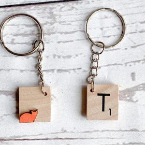 May include: Two keychains featuring square light-colored wooden tiles attached to silver-tone metal rings by short chains. One tile displays a small orange cat silhouette, while the other tile features the black printed letter T with the number 1.