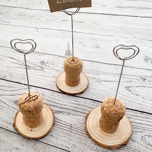 May include: Three cork place card holders with heart-shaped wire holders. The card holders are on a wooden surface. One card holder has a card that says "London".