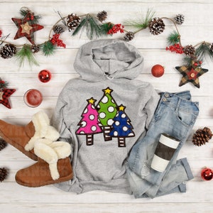 May include: A gray hoodie with a design of three colorful Christmas trees. The hoodie is paired with blue jeans, brown boots, and a coffee cup.