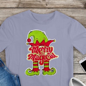 May include: A light purple t-shirt with a green and red elf design. The elf is wearing a green hat with a red and white trim. The text "Merry and Magical" is written in red and yellow letters.