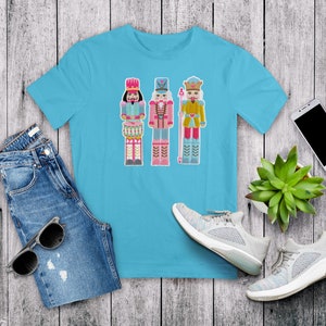 May include: A turquoise blue t-shirt with a graphic of three nutcracker soldiers. The nutcrackers are wearing colorful costumes and holding musical instruments.