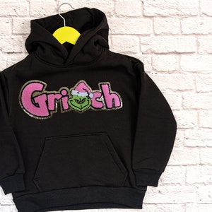 May include: Black hooded sweatshirt with a pink and gold glitter design that says "Grinch" with a green Grinch face in the middle of the word.