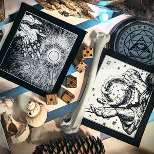Könnte beinhalten: Eine Ansammlung von okkulten Gegenständen, darunter zwei schwarz-weiße Aufnäher mit detaillierten Illustrationen, Holzwürfel, ein Knochen, eine Muschel und ein kreisförmiges Emblem mit einem Augensymbol. Die Gegenstände sind auf einer gemusterten Oberfläche angeordnet.