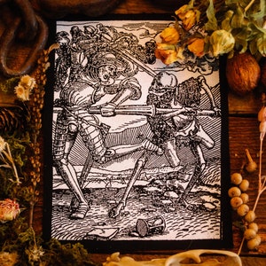 Può includere: Stampa in bianco e nero di un cavaliere che combatte uno scheletro. La stampa è su uno sfondo di tessuto nero. La stampa è circondata da fiori secchi e altri elementi naturali.