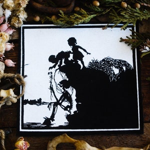 May include: Black and white art print featuring a silhouette of two children on a cliff, with a skeleton below. The artwork has a dark, gothic theme, with a detailed landscape and a stark contrast between light and shadow.