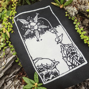 Könnte beinhalten: Schwarz-weiß gestickter Patch mit einer Blume und einem Schädel mit Dornenkrone. Die Blume hat eine sternförmige Mitte und ist von einem Kreis aus Blättern umgeben. Der Schädel ist nach links gerichtet und trägt eine Dornenkrone auf dem Kopf. Der Patch befindet sich auf einem dunklen Hintergrund.