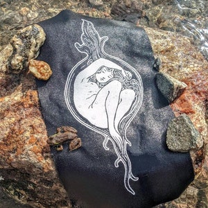 May include: Black fabric with a white illustration of a reclining figure within an ornate, organic frame. The fabric is draped over a large, textured rock. Small rocks and a frog are also visible.