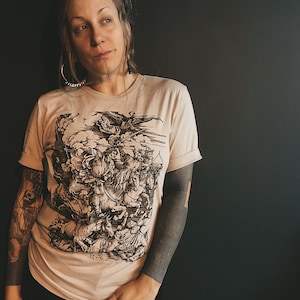 May include: Beige t-shirt with a black and white graphic depicting a historical battle scene. The t-shirt has short sleeves and a crew neck. The person is wearing black trousers.