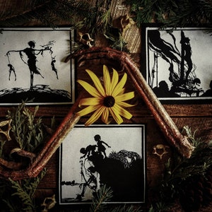 May include: Three square black and white art prints with dark imagery, arranged with a yellow flower, bird foot, and greenery on a wooden surface. The prints depict silhouetted figures in various scenes. The overall aesthetic is dark and artistic.