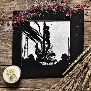 May include: Black and white embroidered patch featuring a silhouette of a figure being burned at the stake. The figure is surrounded by flames and other figures.