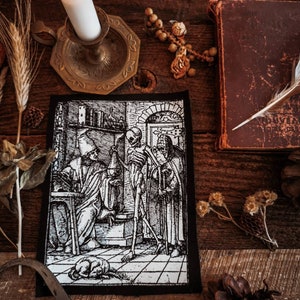 May include: Black and white embroidered patch depicting a scene from "Dance Macabre" by Hans Holbein the Younger. The patch features a skeleton figure holding a scythe, standing next to a man in a robe. A cat is lying on the floor.
