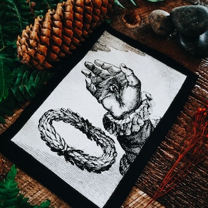 May include: Black and white patch featuring an image of a hand with an eye in the palm, encircled by a wreath of leaves. The patch is displayed on a wooden surface.