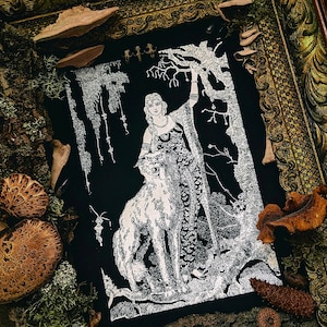 May include: Black and white illustration of a woman with long hair and a decorative dress, standing next to a large wolf. The artwork is framed in a gold-colored ornate frame and surrounded by natural elements like mushrooms and moss.