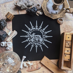 May include: A black square patch featuring a white illustration of a rabbit encircled by swords. The patch is surrounded by dice, bones, a candle holder, and other decorative items on a map.