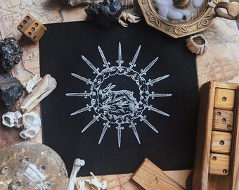 Puede incluir: Un parche cuadrado negro con una ilustración blanca de un conejo rodeado de espadas. El parche está rodeado de dados, huesos, un candelabro y otros elementos decorativos sobre un mapa.