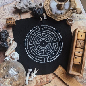 May include: A black square patch with a white labyrinth design. The patch is surrounded by various objects, including dice, a candle holder, a crystal ball, and bones, creating a mystical and esoteric theme.
