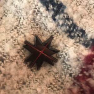 May include: A black star-shaped pin with a red line across the center. The pin is on a white and blue patterned fabric.