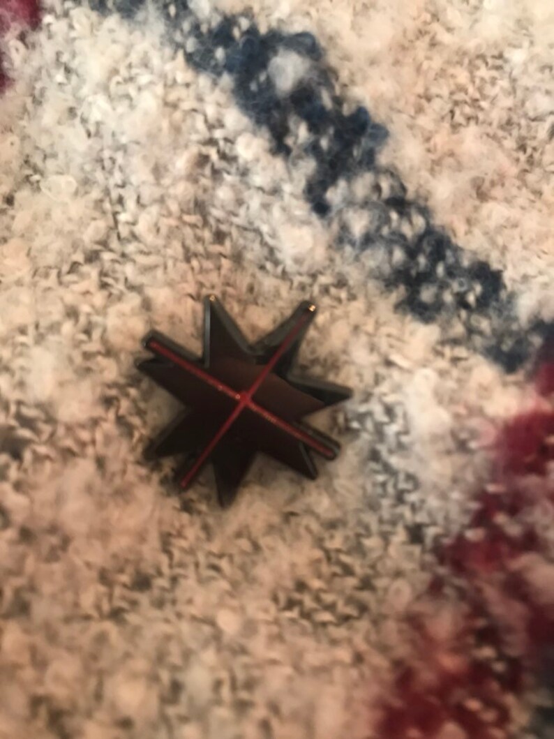 May include: A black star-shaped pin with a red line across the center. The pin is on a white and blue patterned fabric.