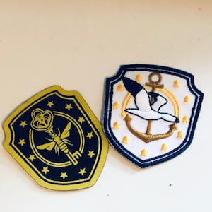 Peut inclure: Deux patchs brodés, l'un avec un motif de clé et d'abeille dorés sur fond bleu marine avec des étoiles dorées, et l'autre avec un oiseau blanc volant au-dessus d'une ancre dorée sur fond blanc avec des étoiles dorées.