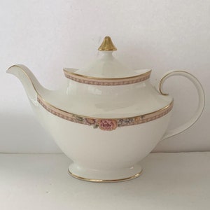 May include: A white porcelain teapot with a gold-coloured finial and trim. The teapot features a decorative band with pink and floral accents. The handle and spout are curved, and the teapot sits on a small foot.