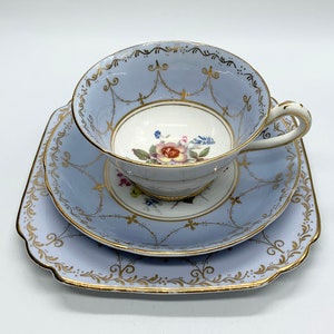 May include: A vintage tea set featuring a light blue teacup, saucer, and square plate. The set has gold trim and a delicate floral design inside the teacup. The teacup is resting on the saucer, which is on top of the plate.