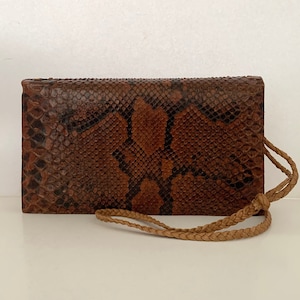 May include: A brown and black snakeskin wallet with a plaited wrist strap. The wallet is rectangular and features a natural pattern. The wrist strap is a light brown colour.