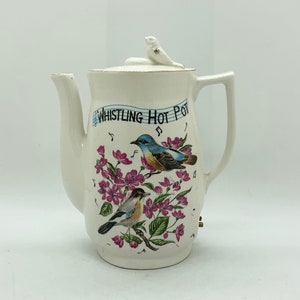 May include: White ceramic teapot with a lid and handle. The teapot is decorated with two birds, pink flowers, and the text "Whistling Hot Pot". A small bird figurine is on the lid.