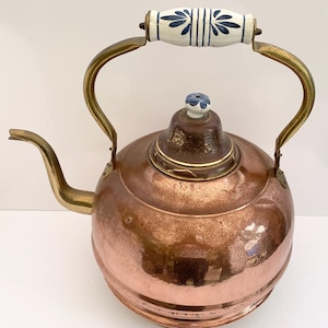 May include: A copper tea kettle with a round body and a curved spout. The handle is made of brass and has a white ceramic grip with blue floral designs. The lid is topped with a small blue flower-shaped knob.