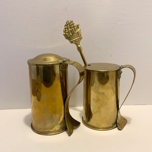 May include: Two brass tankards with lids and handles. A small brass spoon with a ship design rests in one tankard. The tankards have a vintage appearance and are likely decorative or for serving beverages.