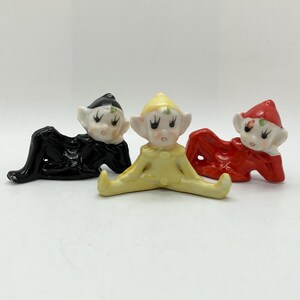 Peut inclure: Trois figurines d'elfes en céramique noire, jaune et rouge. Chaque elfe a un visage blanc avec de grands yeux et des oreilles pointues. Les elfes sont en position couchée, les jambes écartées.