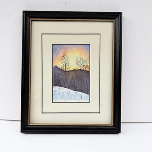May include: A framed watercolour painting depicting a winter landscape with a golden sunset sky, bare trees, and snow-covered ground. The artwork is titled "Hidden Sunlight" and signed by the artist, enclosed in a black frame with a cream-coloured mat.