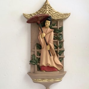 May include: A decorative wall hanging featuring a woman in traditional Asian attire, holding a red parasol. The figure is set against a backdrop of a pagoda-style structure with a trellis and green foliage. The color palette includes beige, gold, red, and green.