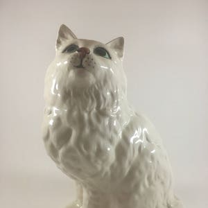 May include: A white ceramic cat figurine with a fluffy coat, green eyes, and a pink nose. The cat is sitting upright, looking upwards. The figurine has a glossy finish and is a decorative item.