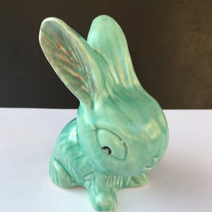 May include: A ceramic bunny figurine in a seated pose, glazed in a soft turquoise color. The bunny has long ears, a small black eye, and a slightly smiling expression. The figurine is a decorative item.