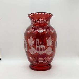 May include: A ruby red glass vase with etched white designs. The vase features a wide base, a cylindrical body, and a flared rim. The etched designs include floral and architectural motifs, with a series of small white dots around the rim.