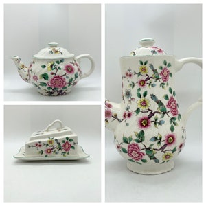 May include: A set of vintage cream-colored ceramic serving pieces with a floral design. The set includes a teapot, a coffee pot, and a covered butter dish. The floral pattern features pink flowers, green leaves, and small birds.