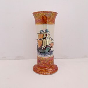 May include: A tall, decorative vase with a flared top and base. The vase features a detailed illustration of a sailing ship on a cream-colored background. The top and bottom sections have a speckled, reddish-brown pattern.