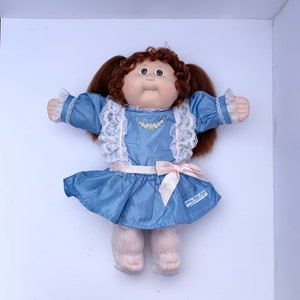May include: A vintage Cabbage Patch Kids doll with brown curly pigtails, wearing a light blue dress with white lace trim and a pink bow. The doll has a soft, fabric body and is lying on a white surface.