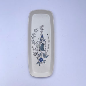 May include: Cream-coloured rectangular ceramic serving tray. It features a whimsical illustration of a figure with a watering can surrounded by blue and grey floral elements. The tray's edges are slightly rounded.